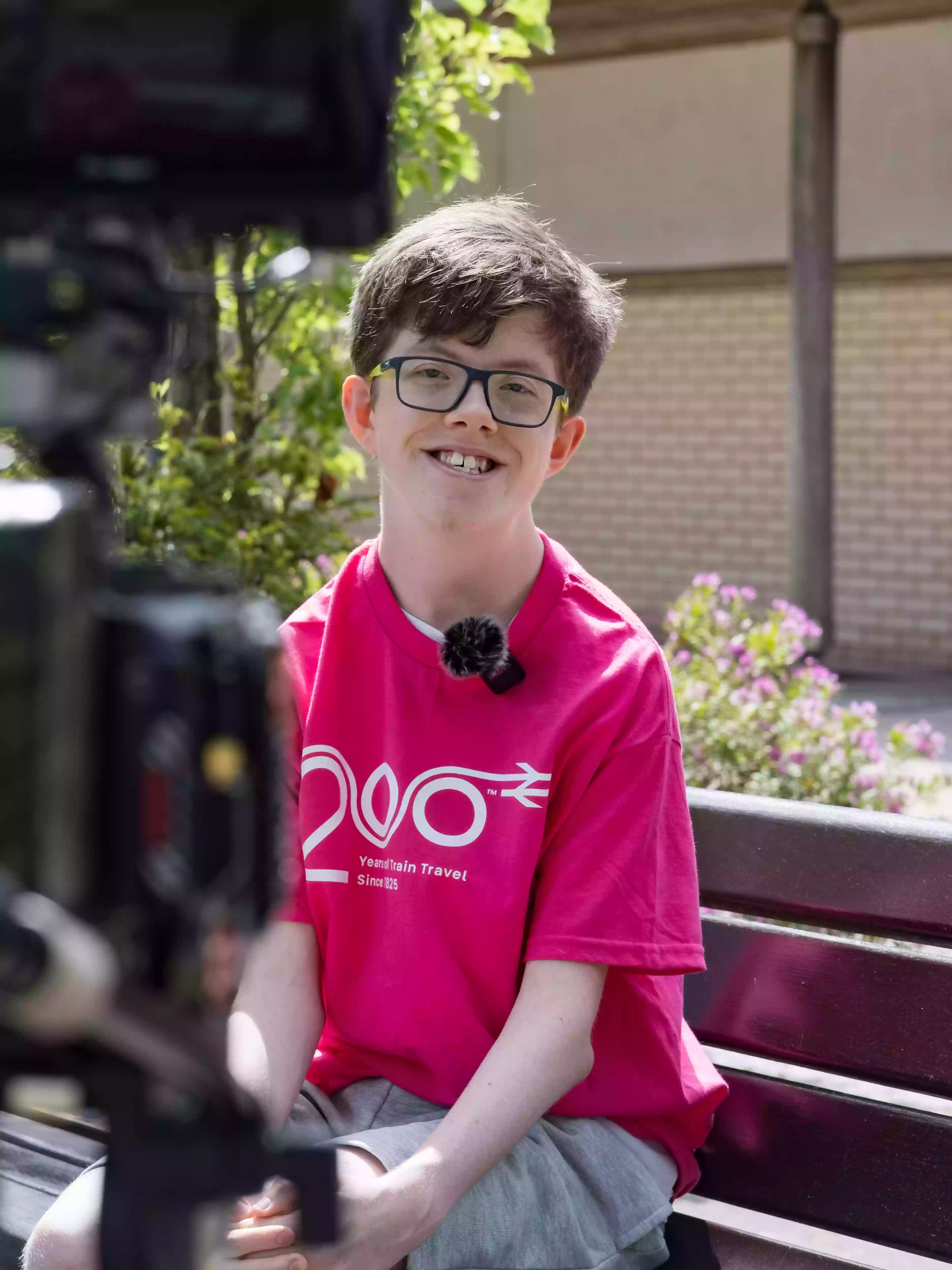 A COPC student wearing a pink Railway 200 t-shirt being filmed for an on-camera interview, with a microphone visible, during behind-the-scenes production coverage