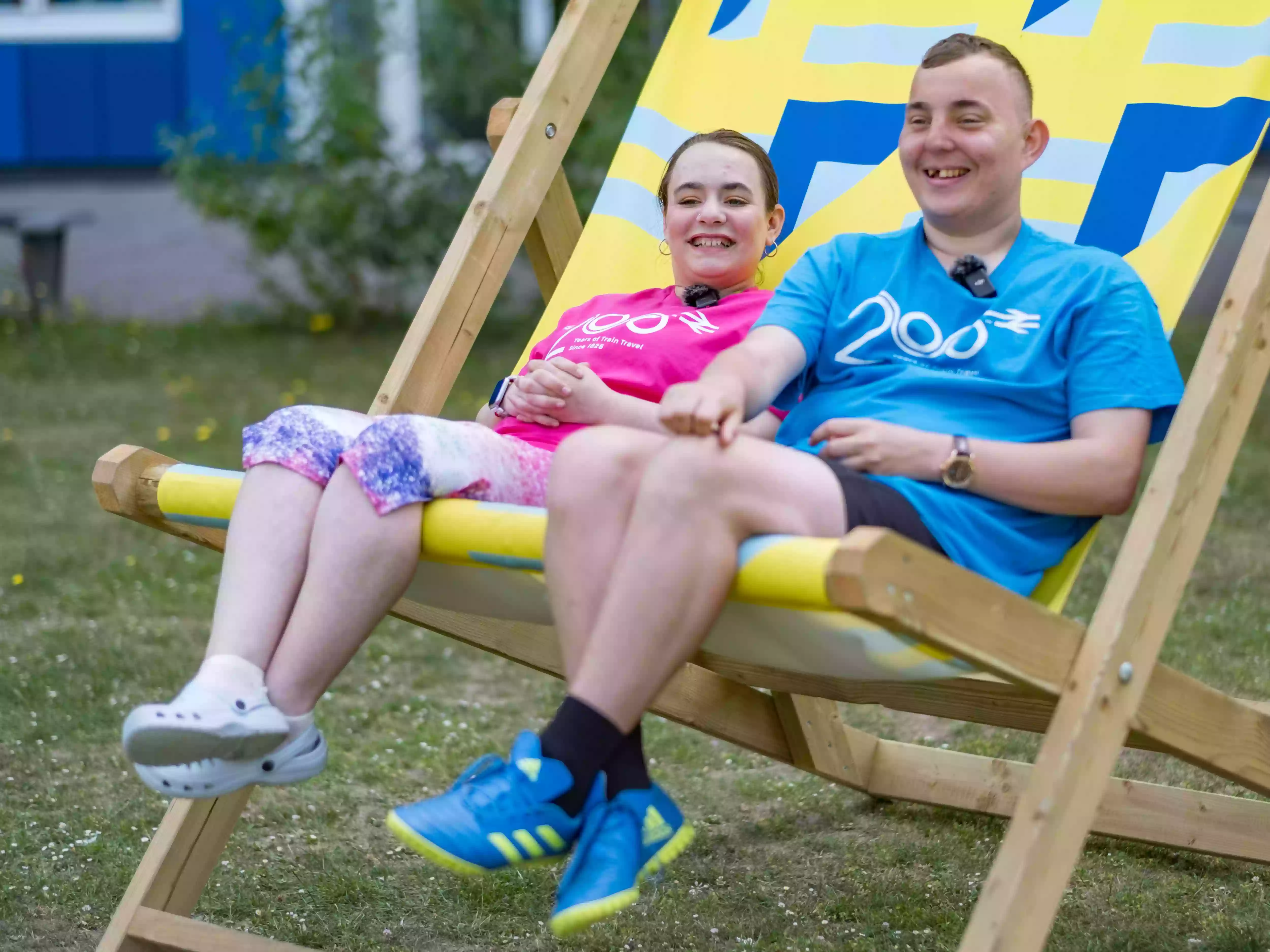 Two COPC students sitting together in a large outdoor deckchair, smiling, one wearing a pink Railway 200 t-shirt, captured during behind-the-scenes filming at the event