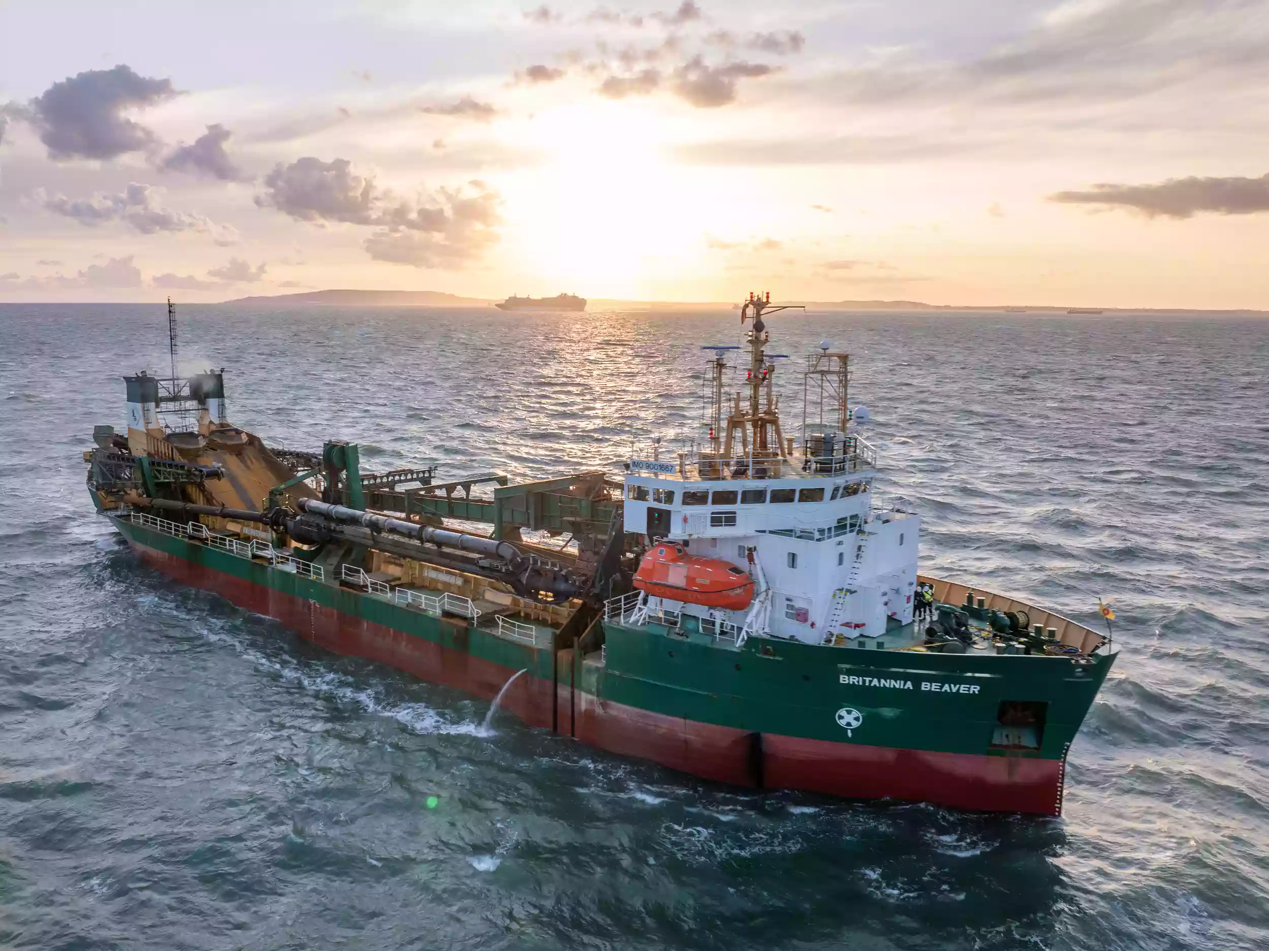 Aerial drone view of the Britannia Beaver dredger at sea, photographed bow-on at sunrise with golden light reflecting on the water.