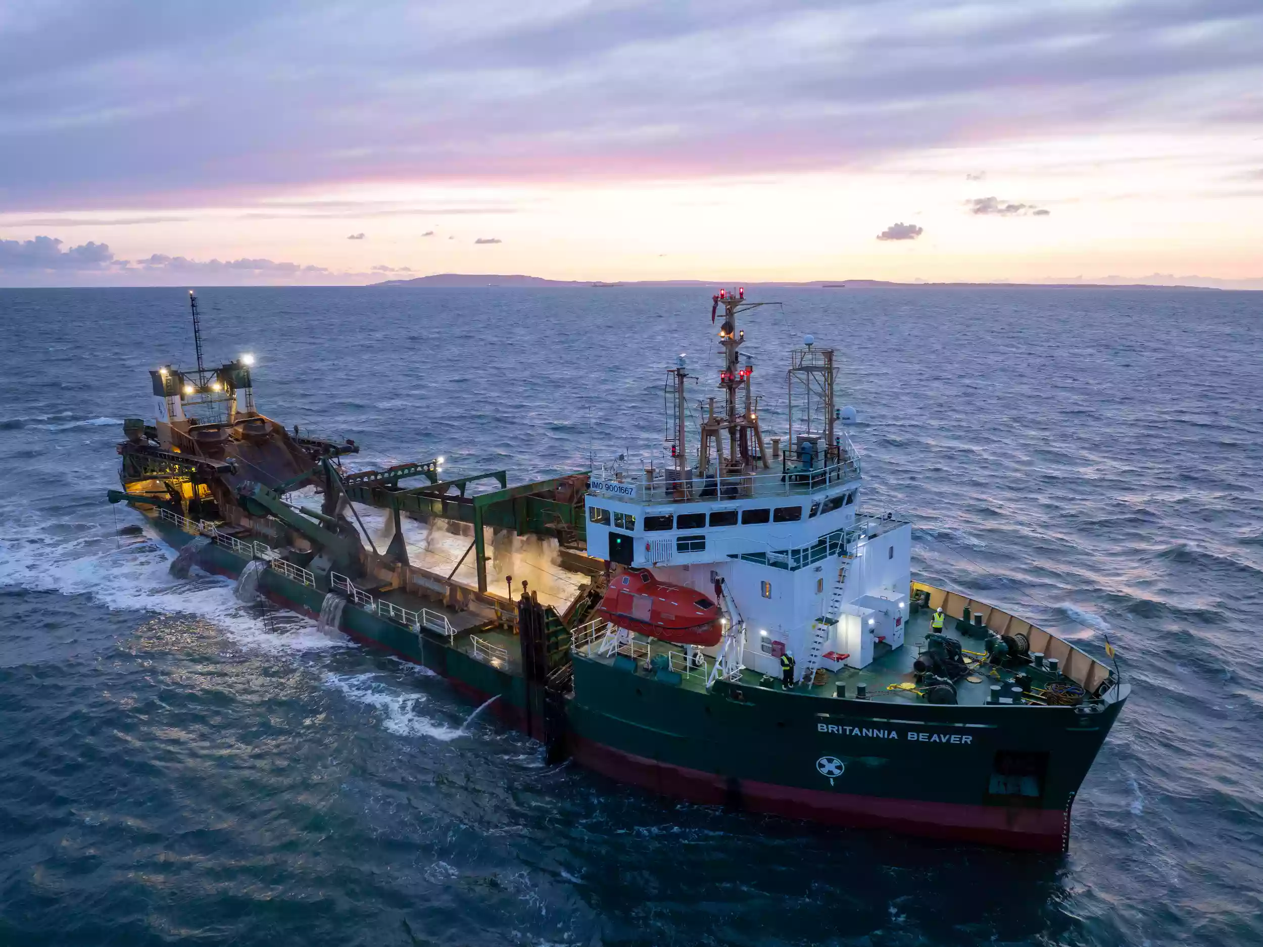 Side profile aerial drone photograph of the Britannia Beaver dredger at sea, taken at dusk with a colourful sky overhead.