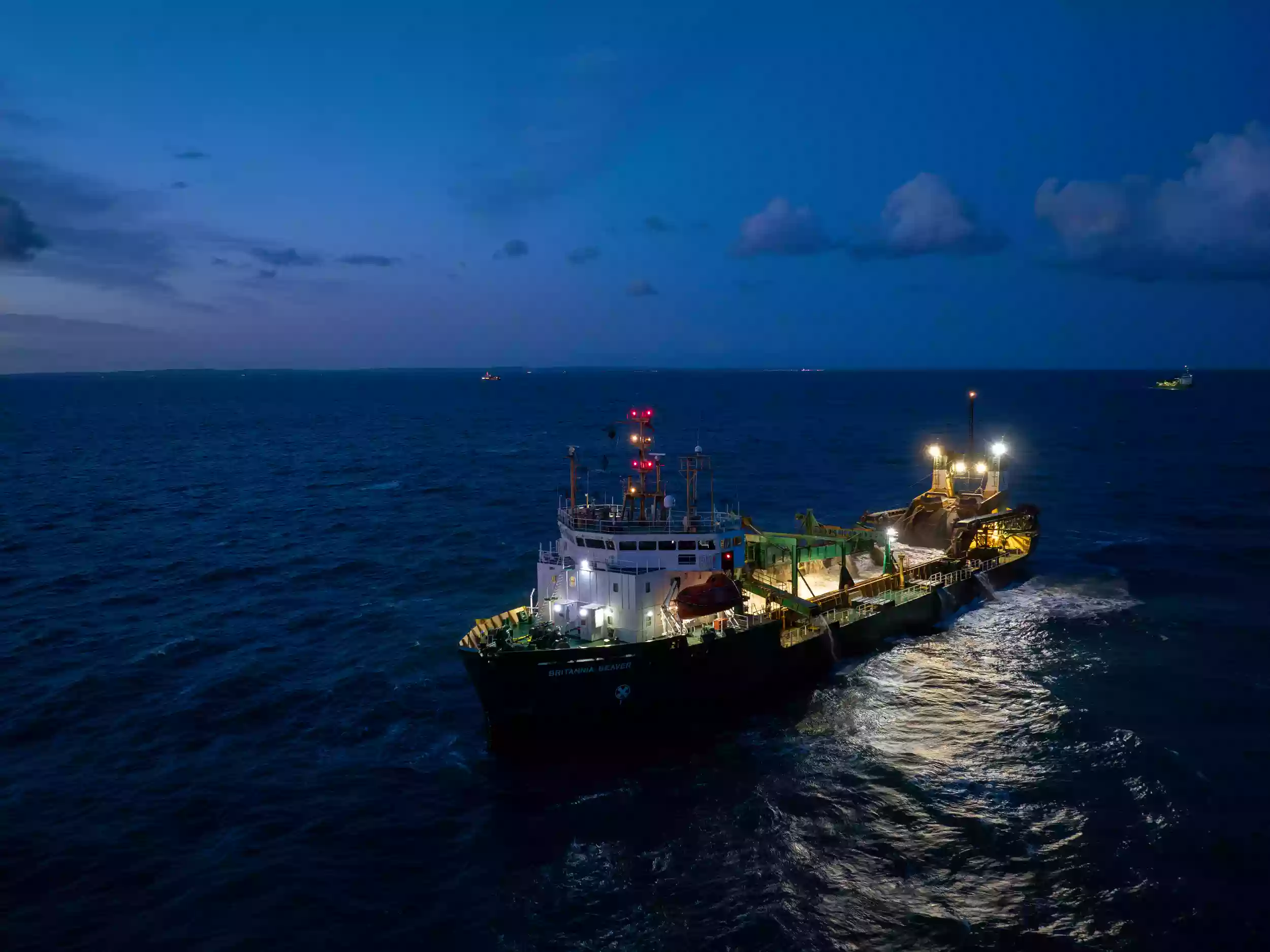 Aerial drone photograph of the Britannia Beaver dredger heading bow-first through open water, with wake trailing behind the vessel.