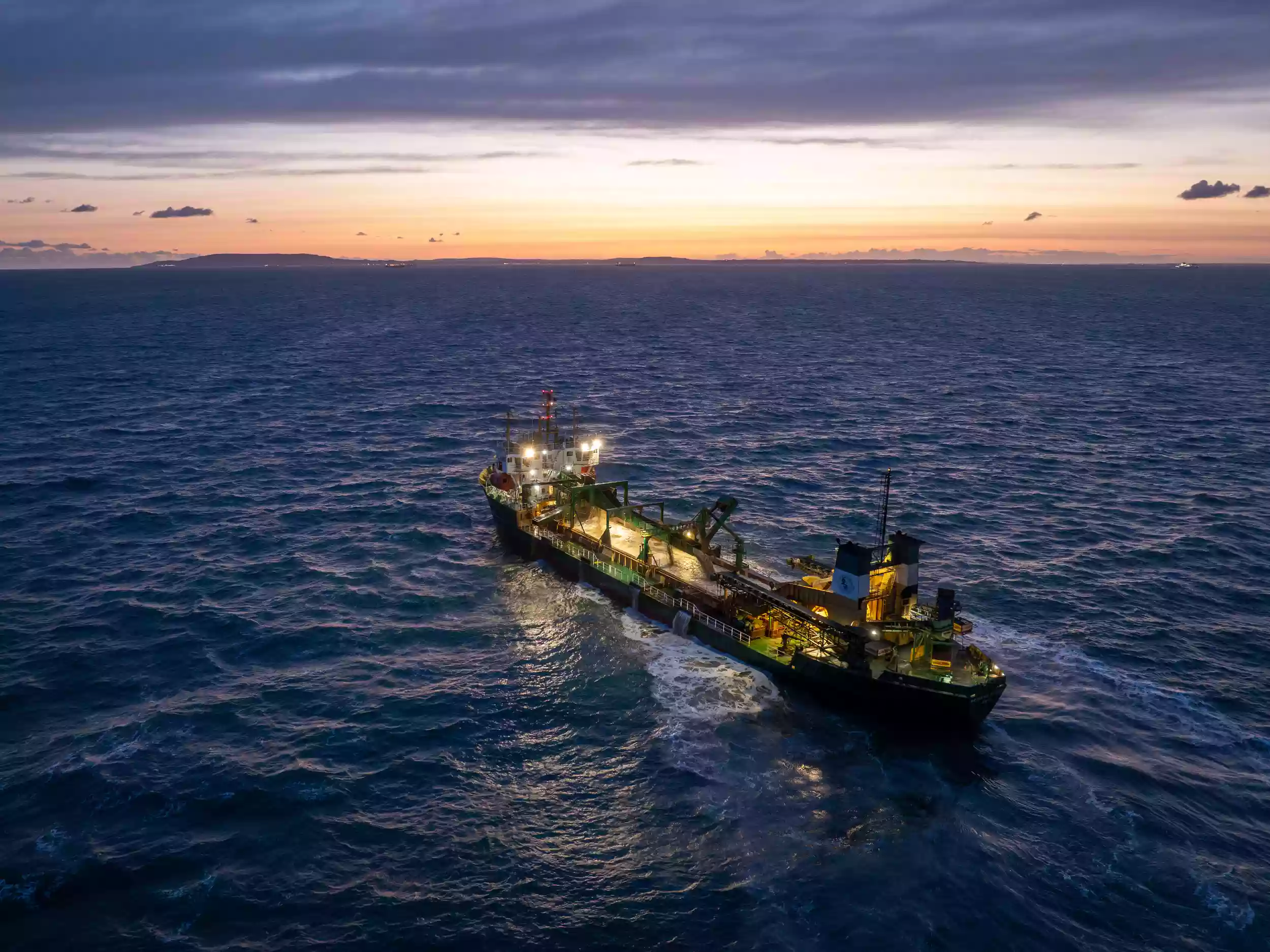 Blue-hour aerial drone shot of the Britannia Beaver at sea with the vessel's deck lights illuminated against a darkening sky.