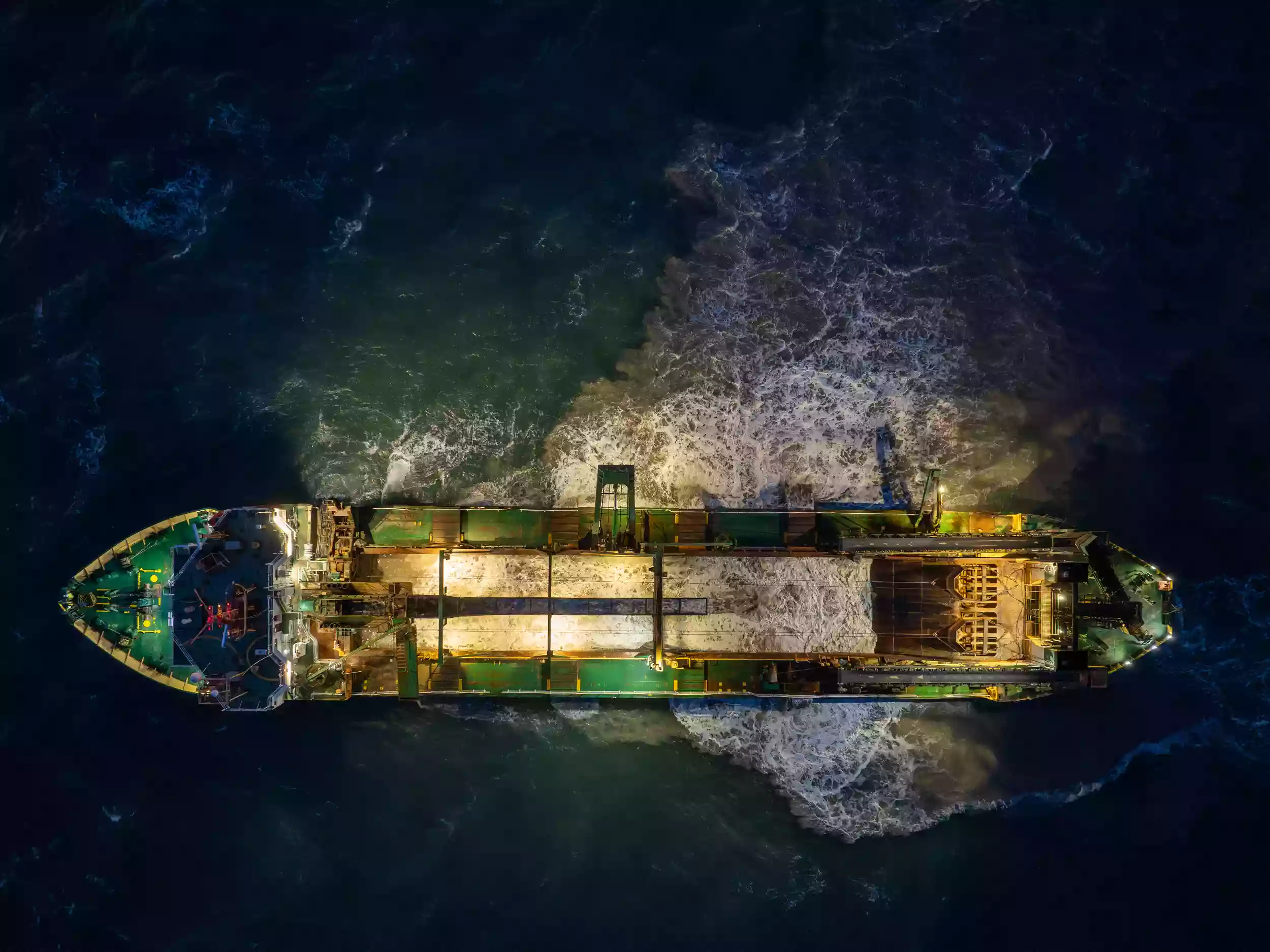 Top-down aerial drone photograph of the Britannia Beaver dredger at night, with the vessel's deck and hopper lit by onboard lighting.