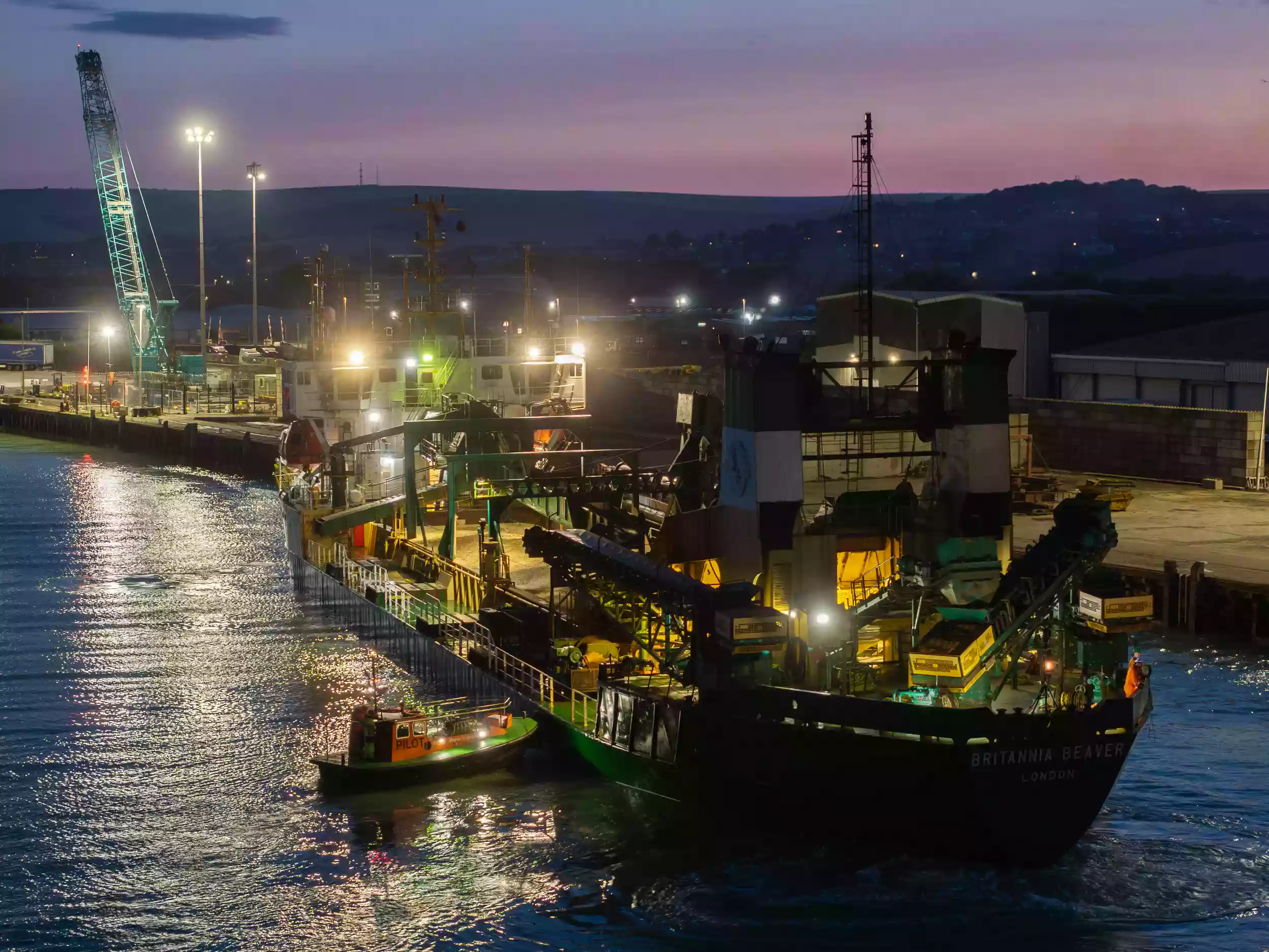 Aerial drone view of the Britannia Beaver berthed in port at night, with the vessel and dockside illuminated against dark surroundings.