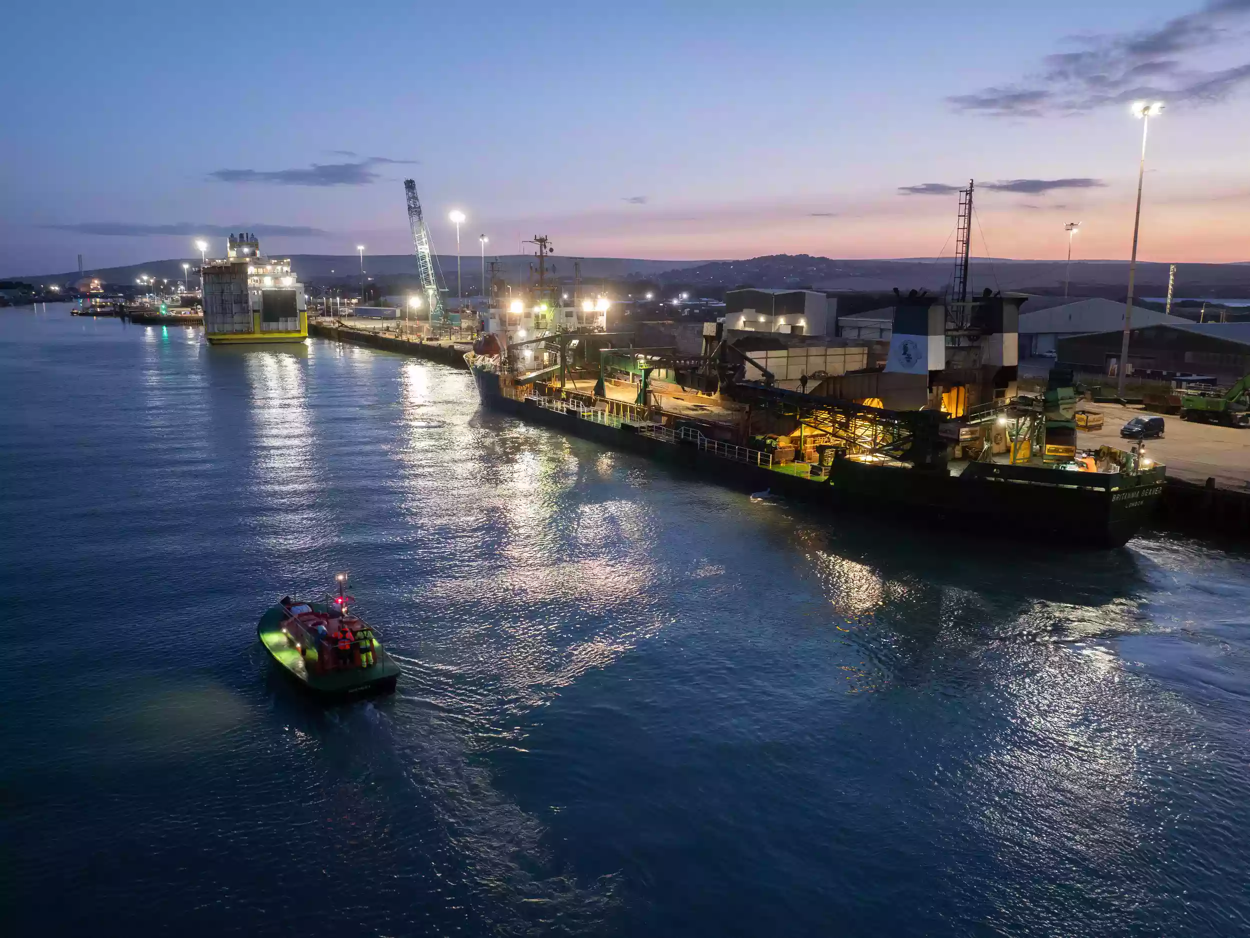 Aerial drone shot of the Britannia Beaver alongside the quay at dusk, with port lights reflecting on the calm harbour water.