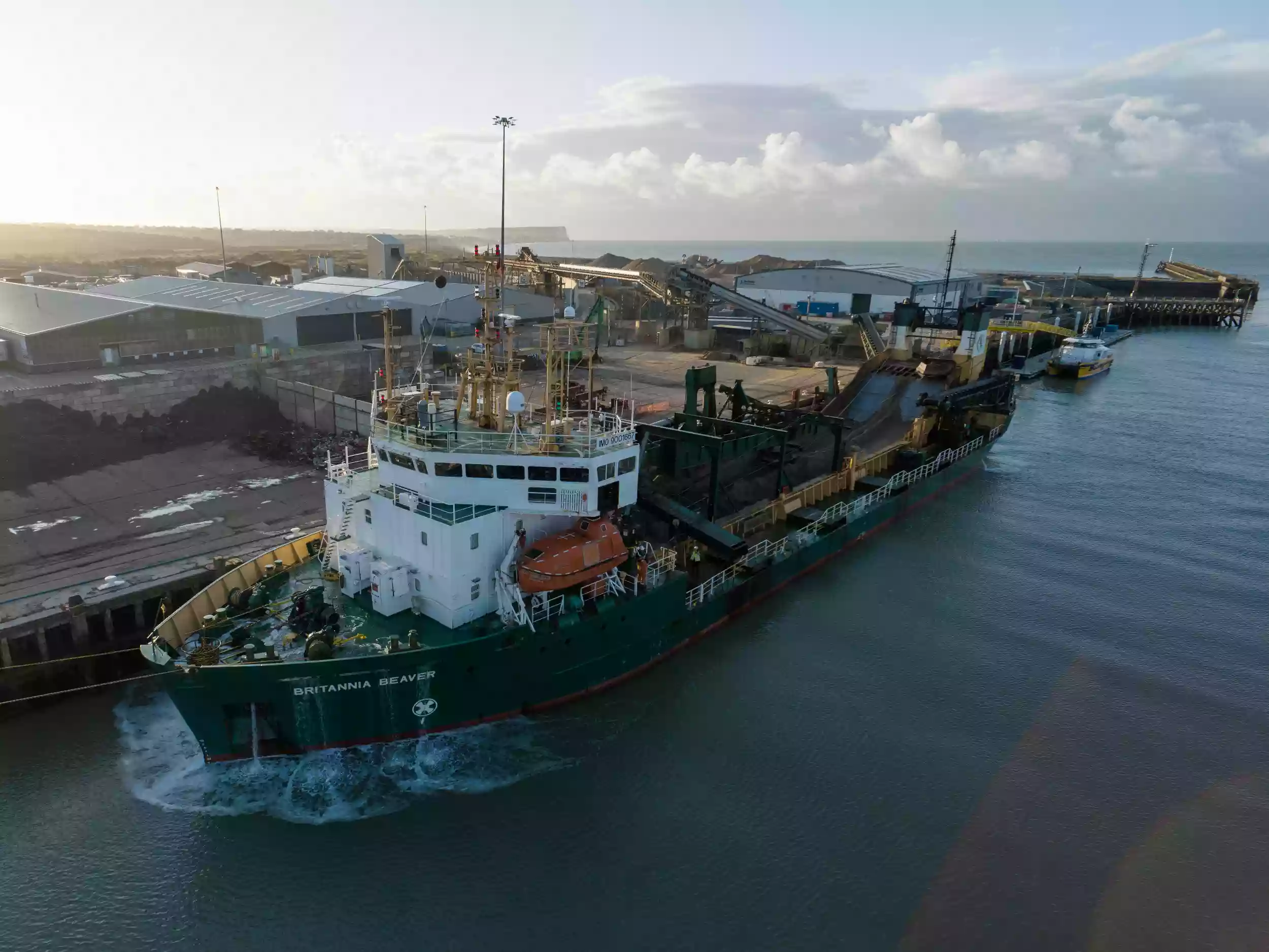 Aerial drone view of the Britannia Beaver moored in an active working port, with cranes and dockside operations visible nearby.