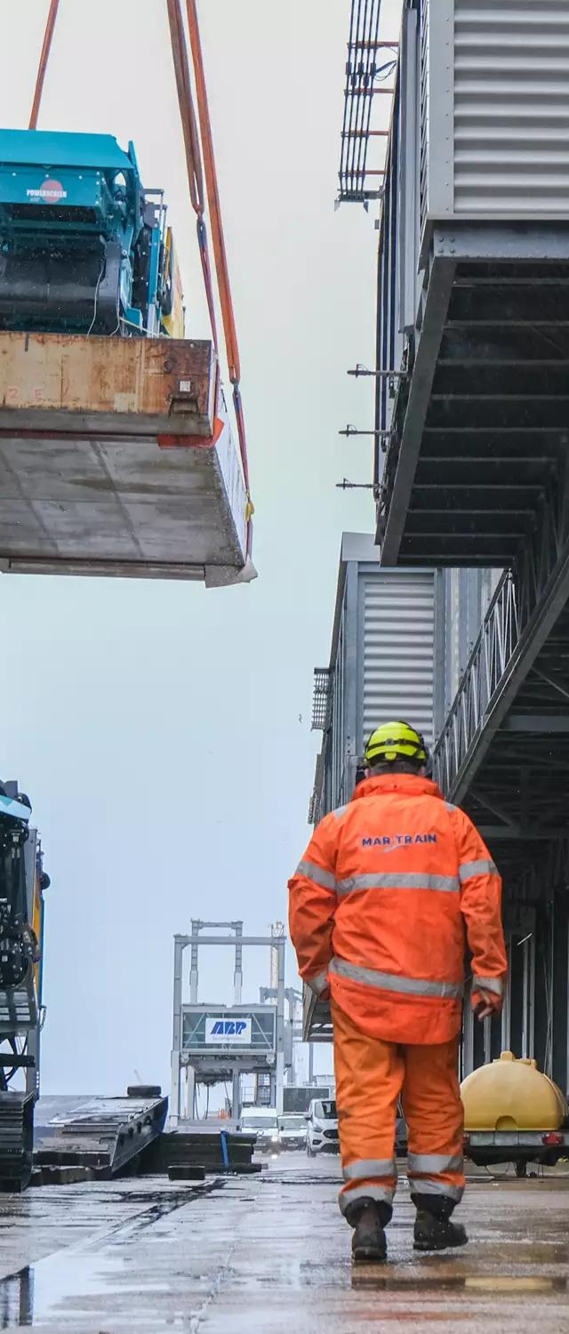 Port worker in orange hi-vis walking the quayside as a Terex crusher is lifted aboard at ABP Southampton
